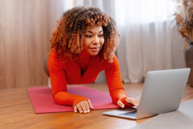 Bir kadın yoga minderinde kalas pozisyonunu koruyor, çevrimiçi fitness programında eğitim görüyor.