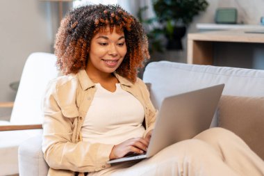 Kadın gülümsüyor, dizüstü bilgisayarda yazıyor, koltukta rahatça oturuyor, uzaktan çalışıyor.