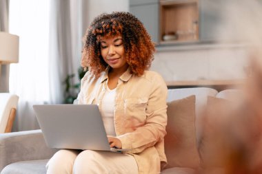 Genç siyahi kadın internette dizüstü bilgisayarla çalışıyor, evdeki kanepede oturuyor, serbest yaşam tarzını temsil ediyor.