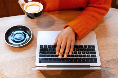 Bir kadının elleri, elinde bir fincan kahve tutarken, tahta bir masanın klavyesinde daktilo kullanıyor.