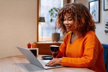 Kıvırcık saçlı, gülümseyen ve dizüstü bilgisayarda yazan bir kadın, evden kahve alıp uzaktan çalışıyor.