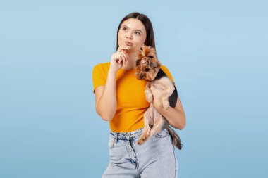 Genç bir kadın, küçük köpeğini mavi bir arka planda tutarak bir düşünce üzerinde kafa yoruyor.