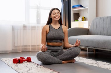Genç bir kadın yoga minderinde lotus pozisyonunda meditasyon yapıyor. Evde zindelik ve sağlık