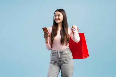 Gülümseyen mutlu ergen, güzel kız alışveriş çantası taşırken akıllı telefon kullanıyor. Alışveriş konsepti