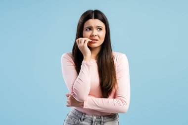 Üzgün, endişeli, tırnak yiyen genç kız stresli, başka tarafa bakıyor, mavi arka planda tek başına duruyor. Sorun, olumsuz duygular kavramı