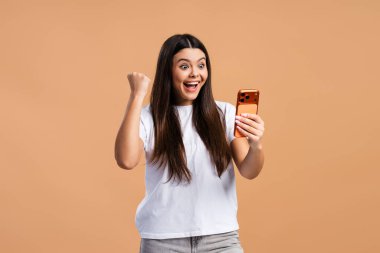 Mutlu, gülümseyen genç kız çevrimiçi başarıyı cep telefonuyla kutluyor. Bej arka planda izole edilmiş para kazanıyor. Zafer konsepti