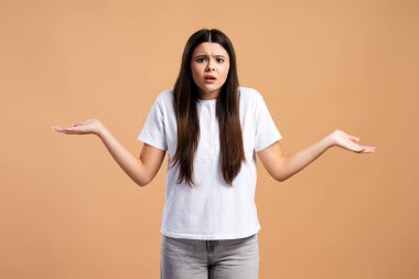Kafası karışmış üzgün bir kız, bej arka planda izole edilmiş tereddütlü bir ifadeyle omuz silkiyor. Duygular konsepti