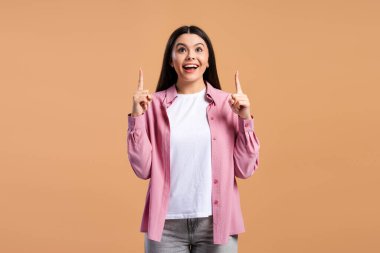 Heyecanlı genç kız iki işaret parmağıyla bej arka planda izole bir şekilde yukarı bakıyor. Reklam kavramı
