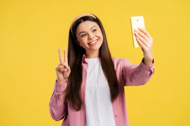 Mutlu gülümseyen genç cep telefonu tutarak sarı arka planda izole edilmiş barış işareti gösteriyor. Çevrimiçi teknoloji kavramı