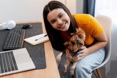 Kendini mutlu hisseden bir kadın, küçük köpeğini dizüstü bilgisayarı, klavyesi ve kulaklığı olan bir masanın yanında tutuyor.