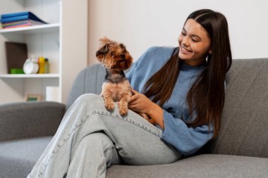 Kadın gülümser, küçük Yorkshire teriyeri köpeğini kucağında kanepede okşar.