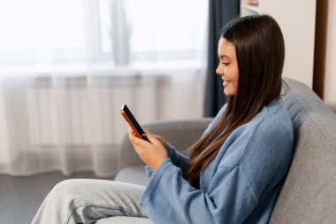 Kanepede oturan genç bir kadın iletişim ve bağlantı kurmak için cep telefonunu mutlulukla kullanıyor.