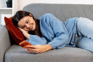 Genç bir kadın gülümsüyor, kanepede uzanıyor, sosyal medya ve rahatlama için akıllı telefon kullanıyor.