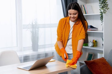 Masayı sprey şişesi ve bezle temizleyen genç bir kadın, temiz bir ev ortamı sağlıyor.