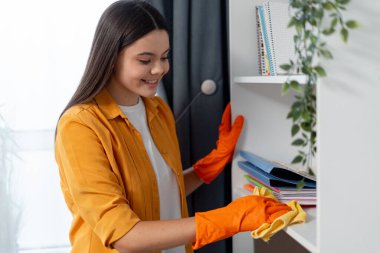 Turuncu eldiven giyen genç bir kadın, evinde beyaz bir rafı temizleyip tozluyor.