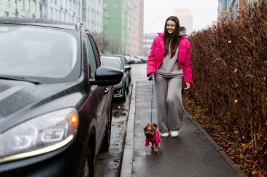 Pembe ceketli genç bir kadın modern şehir ortamında sevimli köpeğini gezdiriyor.