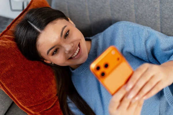 Genç bir kadın gülümsüyor, kanepede uzanıyor, akıllı telefon kullanıyor ve evde internet bağlantısı kuruyor.