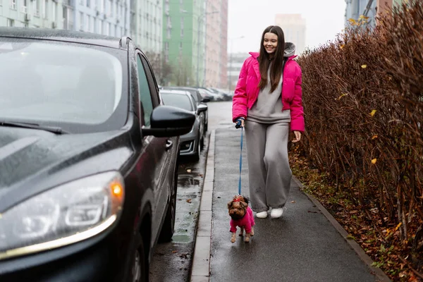 Pembe ceketli genç bir kadın modern şehir ortamında sevimli köpeğini gezdiriyor.