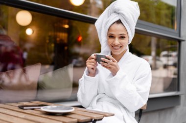 Beyaz bornoz ve havlu giymiş, açık kafe masasında kahve içen, spa sonrası rahatlama ve kendine bakmanın tadını çıkaran genç bir kadın. Rahatlatıcı kavram