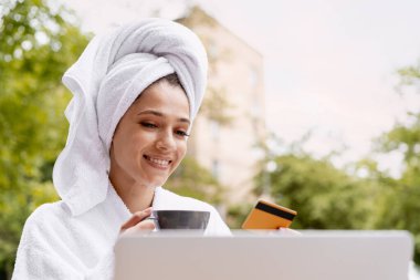 Kafasında beyaz bornoz ve havlu olan güzel bir kadın, dizüstü bilgisayar kullanırken elinde kahve fincanı ve kredi kartı tutuyor. Çevrimiçi alışveriş kavramı