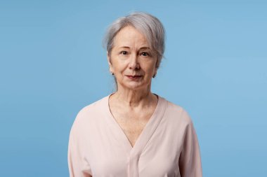 Stüdyo resmi için poz veren yaşlı bir kadın, mavi arka planda doğal yaşlanma ve olgunluğu gösteriyor.