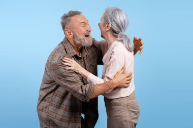 Yaşlı adam ve kadın sıcacık sarılıyor, açık mavi bir arka planda neşeyle gülüyorlar.