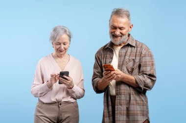 Ayakta duran yaşlı adam ve kadın, teknolojiyle bağlantı kuruyor, mutlu bir şekilde cep telefonu kullanıyor.