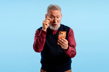 Yaşlı adam şaşkınlıkla tepki veriyor, elinde akıllı telefon ve mavi arka planda gözlük tutuyor.