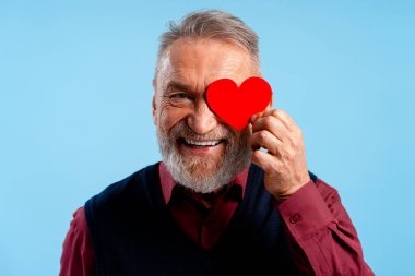 Kırmızı bir kalbi gözünün üstünde tutarken gülümseyen mutlu kıdemli adam, sevgi ve neşe ifade ediyor.