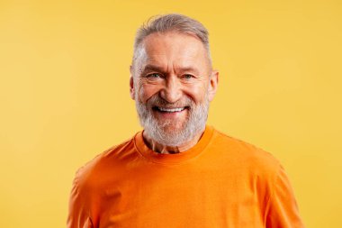 Beyaz sakallı, sarı bir stüdyoda gülümseyen ve kameraya bakan yetişkin bir adam.