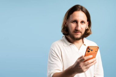 Mavi arka plandaki kameraya bakarken elinde turuncu akıllı telefon tutan genç adam.