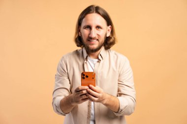 Genç adam gülümsüyor, elinde turuncu bir akıllı telefon, cep telefonu teknolojisi ve internet iletişimi ile meşgul.