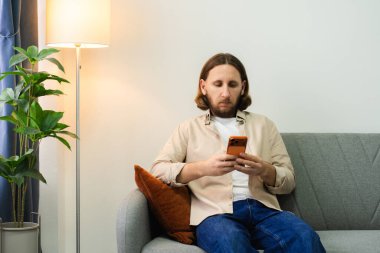 Oturma odasında akıllı telefon kullanan, çevrimiçi bağlantı kuran ve içeriği rastgele kaydıran bir adam