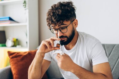 Üzgün, hasta adam soğuk alerji rhinitis tedavisi için burun spreyi kullanıyor evde, kanepede oturuyor. Erkek hasta üşütmüş. Sağlık hizmeti kavramı