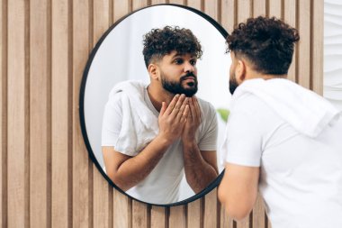 Evdeki banyo aynasında sakalı inceleyen yakışıklı bir ortadoğulu. Sabah rutini konsepti