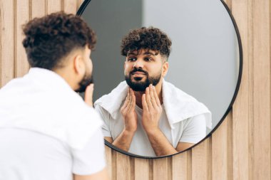 Sakallı adam tıraş losyonu sürüyor, günlük sakal tımar rutini uyguluyor aynaya bakıyor. Sabah rutini konsepti