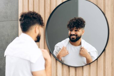 Banyo aynasında gülümseyen kendinden emin orta doğulu adam modern banyodaki özgüvenini yansıtıyor. Sabah rutini konsepti