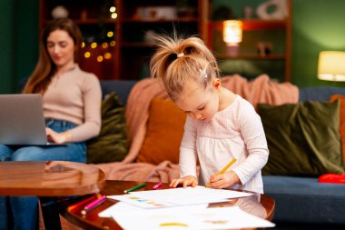 Anne kanepede uzaktan çalışırken çocuk kahve masasına resim çiziyor.