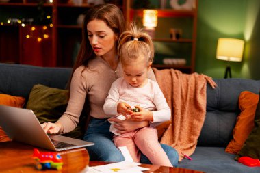 Annem evde dizüstü bilgisayarla çalışıyor, kucağında bebekle kanepede oynuyor.