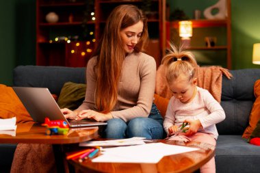 Anne dizüstü bilgisayara odaklanırken kız kanepede yanında kağıt işçiliği yapıyor.