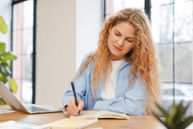 Ciddi bir kadın dizüstü bilgisayarını ve web sitesini kullanarak not defterine not alıyor. Çevrimiçi iletişim kavramı