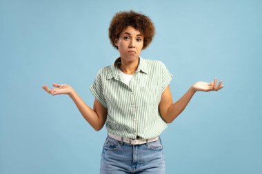 Kafası karışmış Afrikalı Amerikalı kadın kollarını silkip mavi arka planda kafa karışıklığı ve belirsizlik gösteriyor. Sorun kavramı