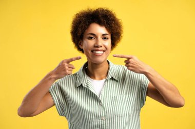 Genç gülümseyen Afro-Amerikan kadın kusursuz beyaz dişlerini göstererek sarı arka planda izole edilmiş kameraya bakıyor. Diş sağlığı konsepti