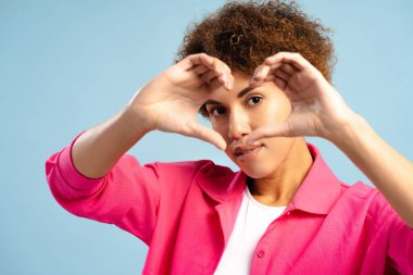 Güzel Afro-Amerikan kadın kalp hareketi yapıyor. Sevgisini ve sevgisini ifade ediyor. Mavi arka planda izole edilmiş kameraya bakıyor. Aşk, duygu konsepti
