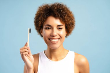 Çevre dostu bambu diş fırçası tutan mutlu gülümseyen Afrikalı Amerikalı kadın mavi arka planda izole bir şekilde poz veren kameraya bakıyor. Diş sağlığı konsepti