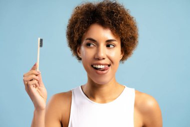 Elinde diş fırçası tutan, ağız temizliğini teşvik eden, mavi arka planda izole edilmiş kameraya bakan Afro-Amerikan kadının portresi. Diş hekimi kavramı