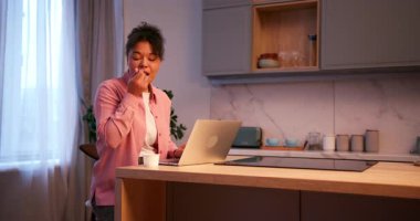 Young woman working on a laptop computer and having a healthy snack in her modern kitchen at home
