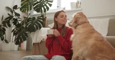 Genç bir kadın, sıcak bir oturma odasında bir kupadan içerken av köpeğini okşuyor.