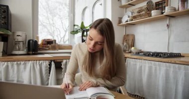 Mutfak masasından internetten öğrenirken not alan odaklanmış kız öğrenci.