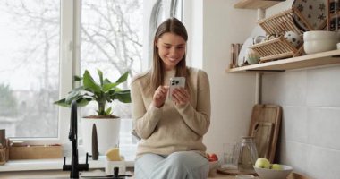 Mutfak tezgahında oturan mutlu kadın, cep telefonuyla mesaj atıyor ve kaydırıyor.